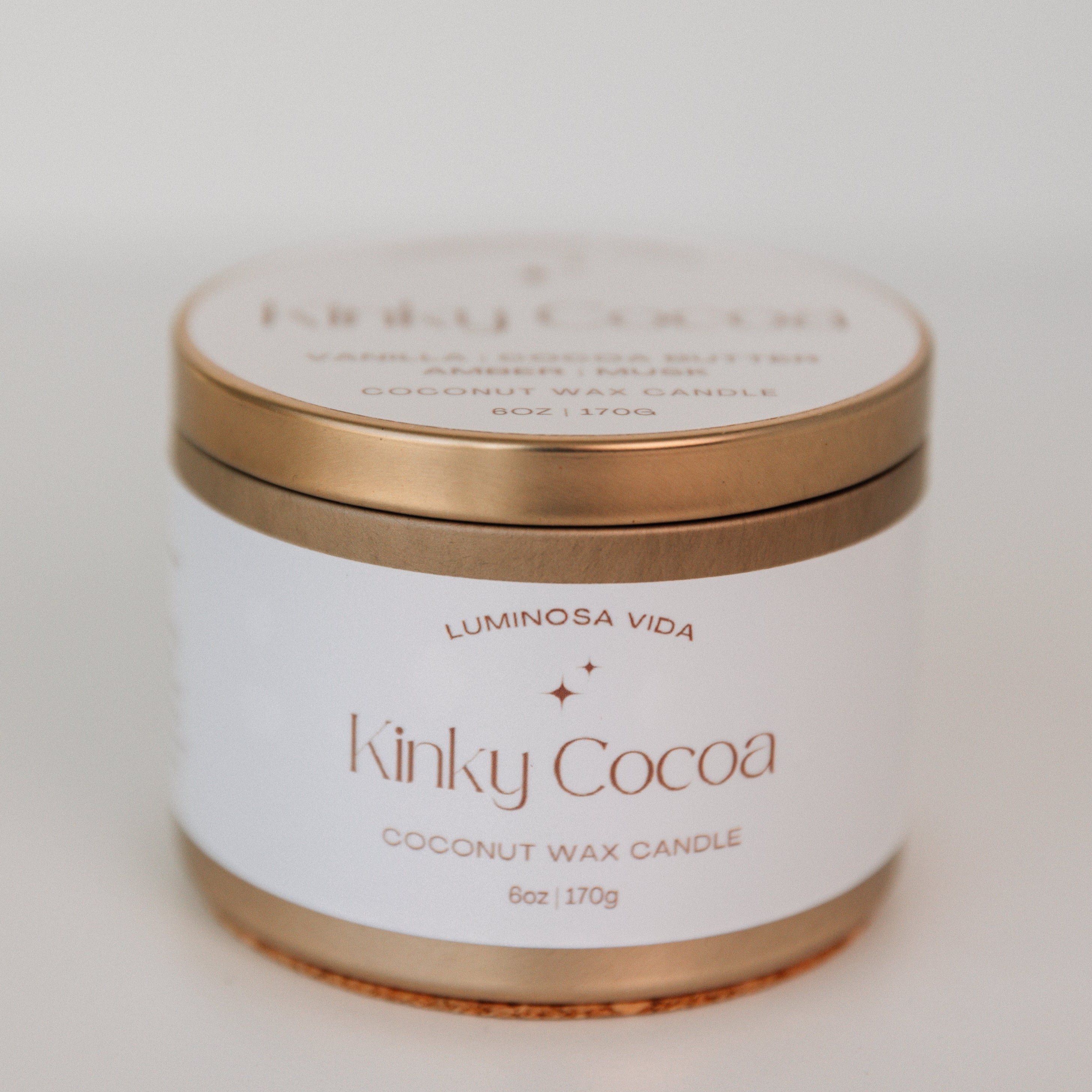 Close-up of a luxurious candle labeled 'Kinky Cocoa,' featuring a rich chocolate-brown wax with a sleek gold accent. The scene is styled with warm cocoa tones, a hint of cinnamon sticks, and a cozy background evoking indulgence and sensuality