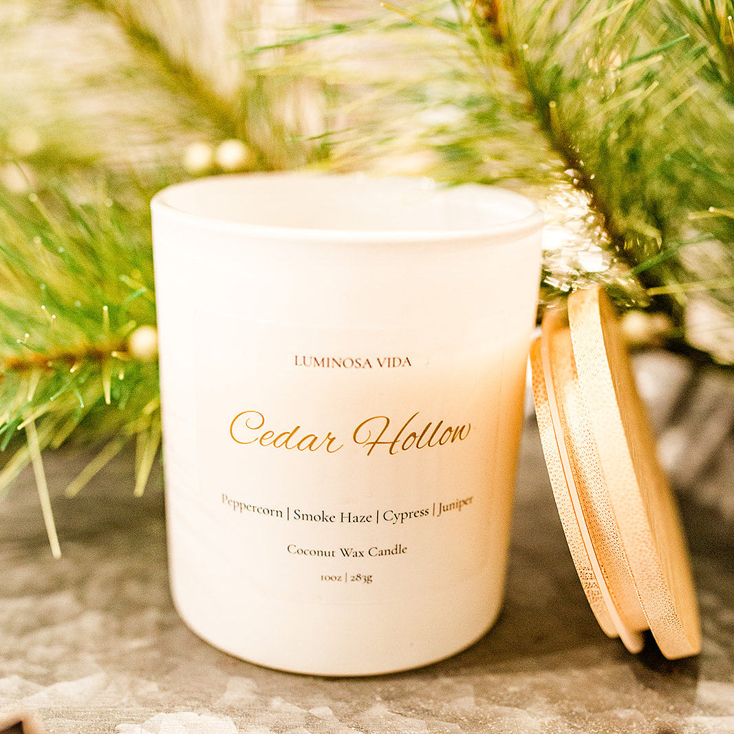“Cedar Hollow candle by Luminosa Vida displayed on a decorative pedestal with festive pine greenery. White jar with gold lid featuring notes of peppercorn, smoke haze, cypress, and juniper.”