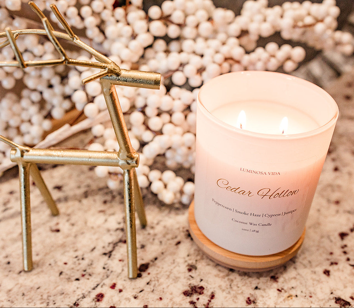 “Lit Cedar Hollow candle by Luminosa Vida displayed on a granite surface beside a gold reindeer figurine and snowy holiday berries. White coconut wax candle featuring notes of peppercorn, smoke haze, cypress, and juniper.”