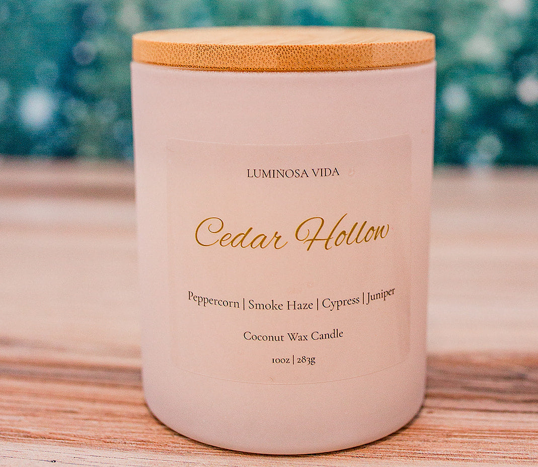 “Cedar Hollow candle by Luminosa Vida on a wooden surface with a winter forest background. White jar with a bamboo lid featuring notes of peppercorn, smoke haze, cypress, and juniper.”