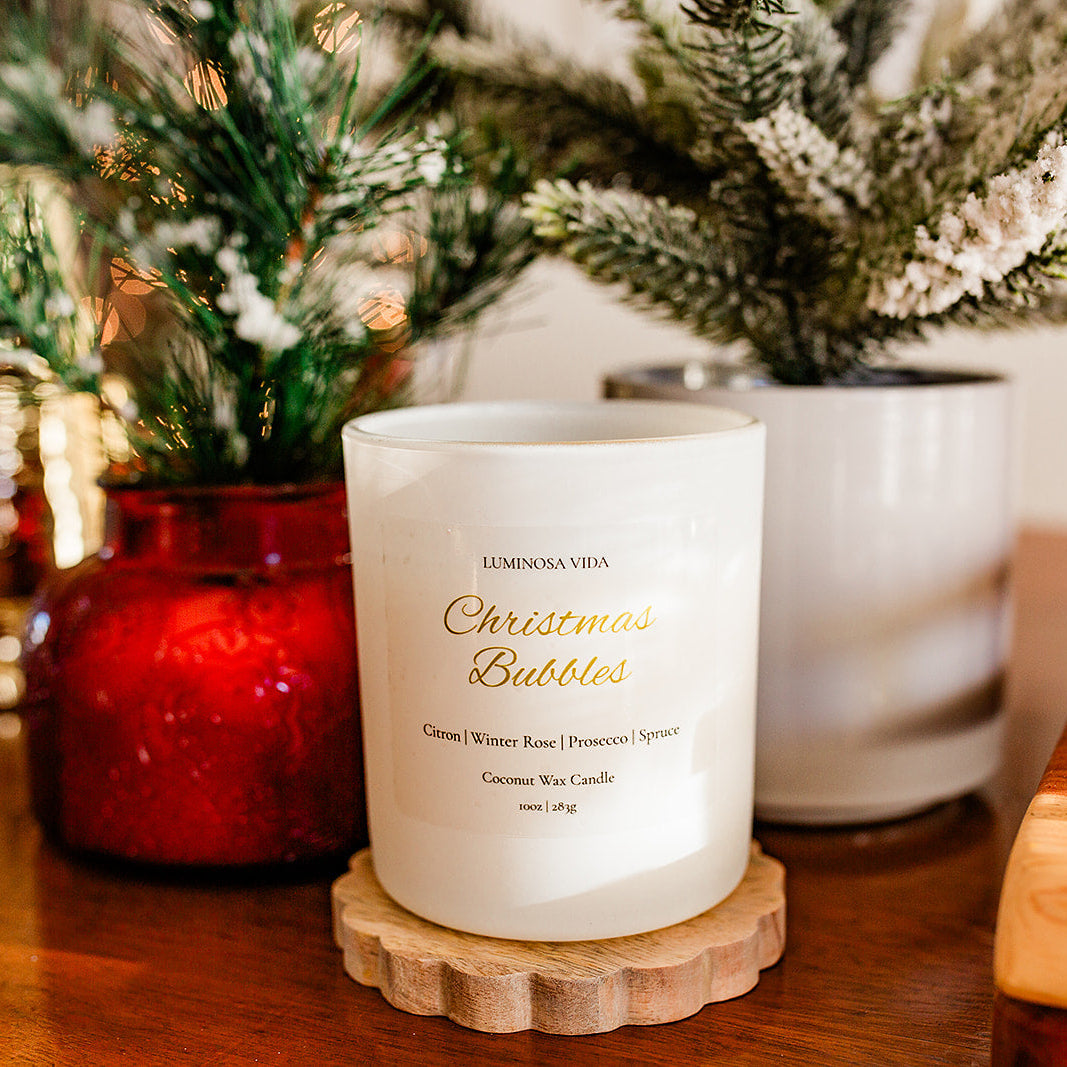 “Christmas Bubbles holiday candle by Luminosa Vida displayed on a wooden tray beside festive décor, including a red glass vase and snowy evergreen branches. White candle jar with gold lettering featuring notes of citron, winter rose, prosecco, and spruce.”