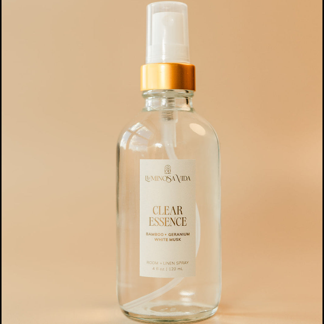 Luminosa Vida Clear Essence room and linen spray with bamboo, geranium, and white musk in a clear glass bottle with gold collar on a neutral background.