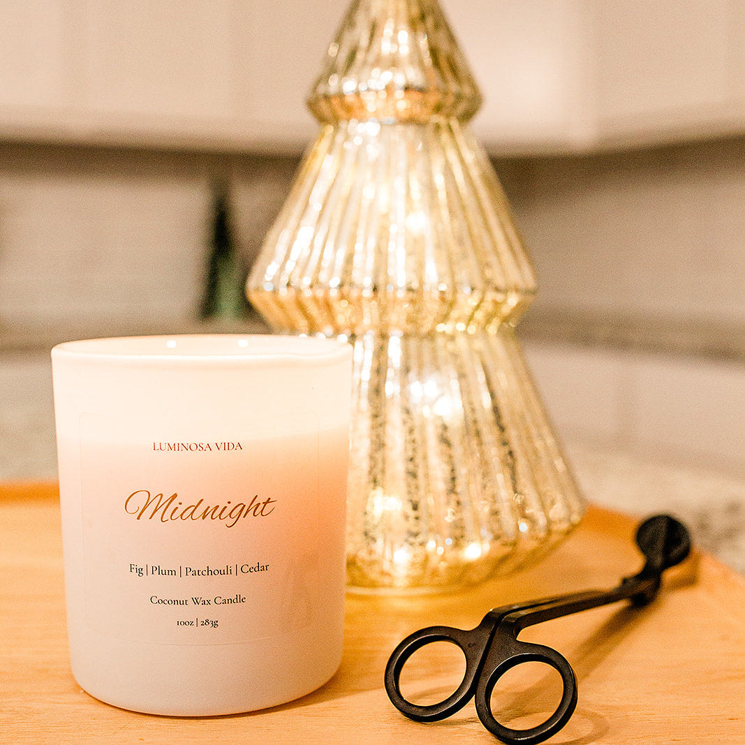 “Midnight candle by Luminosa Vida displayed on a wooden tray beside a gold mercury glass Christmas tree and black wick trimmer. White coconut wax candle featuring notes of fig, plum, patchouli, and cedar.”