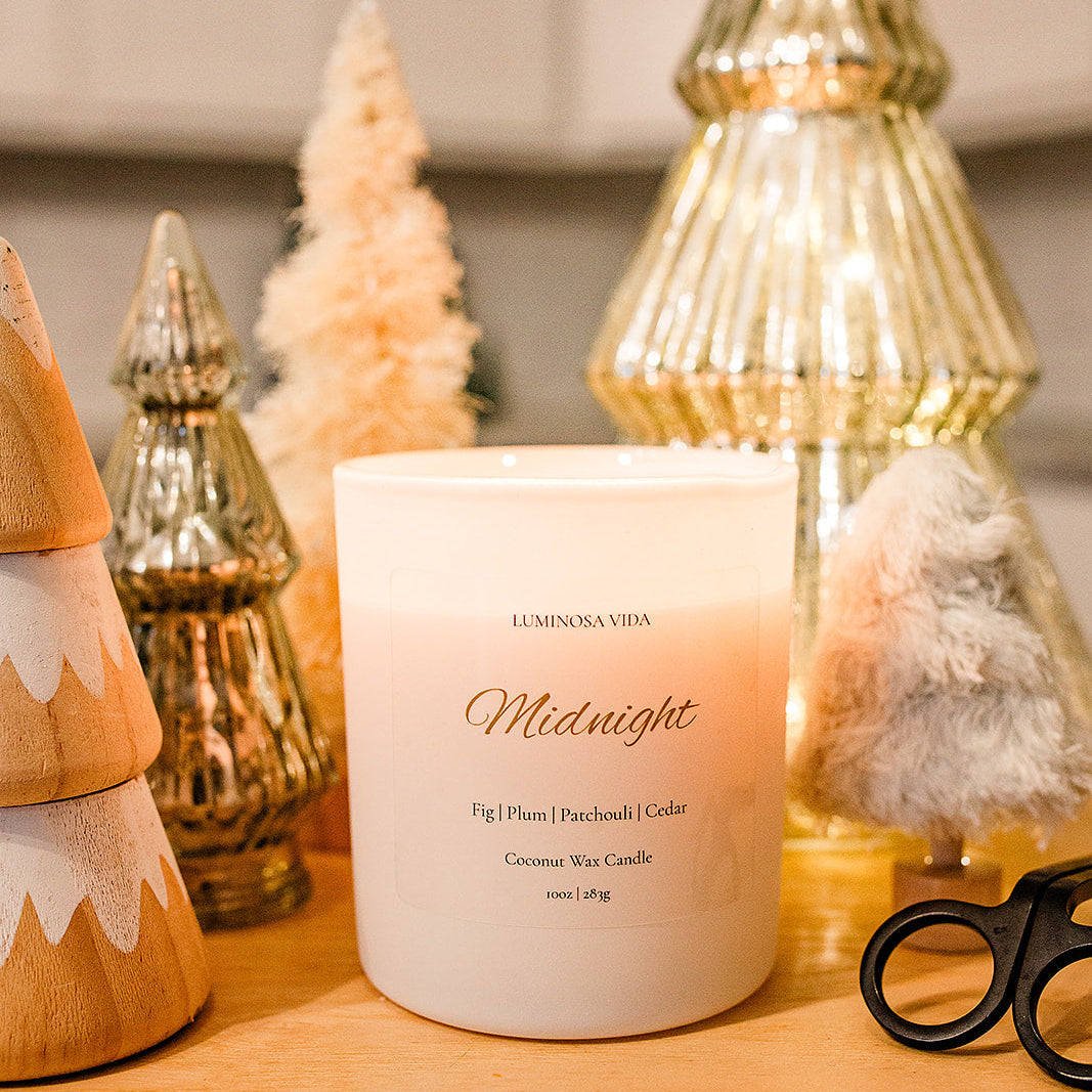 “Lit Midnight candle by Luminosa Vida surrounded by decorative holiday trees in gold, wood, and bottle-brush textures. White coconut wax candle featuring notes of fig, plum, patchouli, and cedar.”