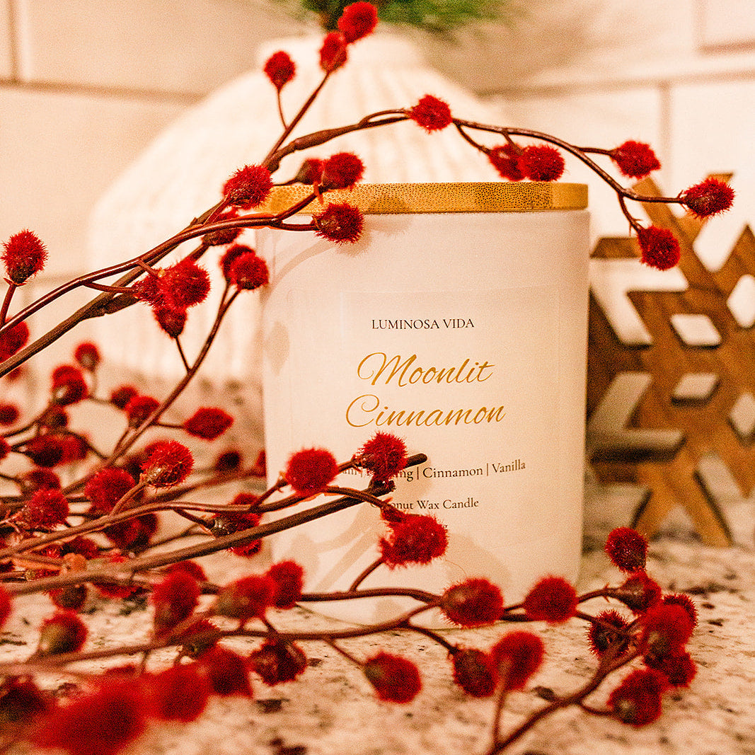 “Moonlit Cinnamon candle by Luminosa Vida surrounded by red holiday berry stems and winter greenery on a speckled countertop. White coconut wax candle featuring notes of cream, pudding, cinnamon, and vanilla.