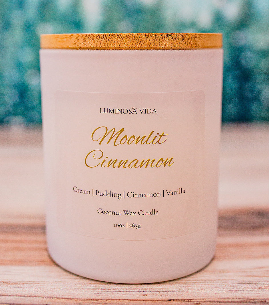 Moonlit Cinnamon candle by Luminosa Vida on a wooden surface with a snowy forest backdrop. White jar with bamboo lid featuring notes of cream, pudding, cinnamon, and vanilla.