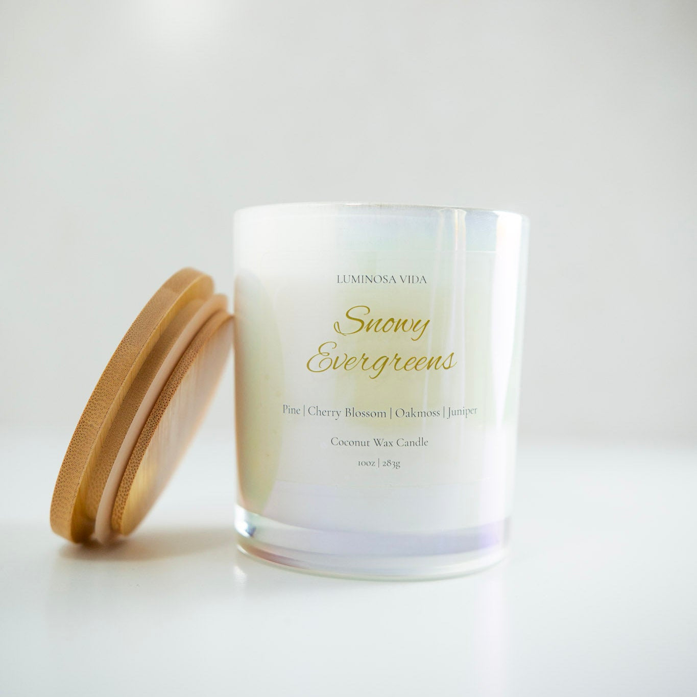 “Luminosa Vida Snowy Evergreens candle in an iridescent glass jar with a wooden lid. Hand-poured coconut wax candle made in Austin, Texas with notes of pine, cherry blossom, oakmoss, and juniper — a fresh winter fragrance.”