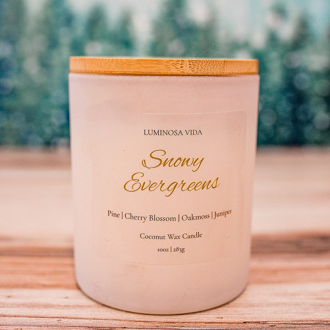 “Snowy Evergreens candle by Luminosa Vida on a wooden surface with a soft winter forest background. White jar with bamboo lid featuring notes of pine, cherry blossom, oakmoss, and juniper.”