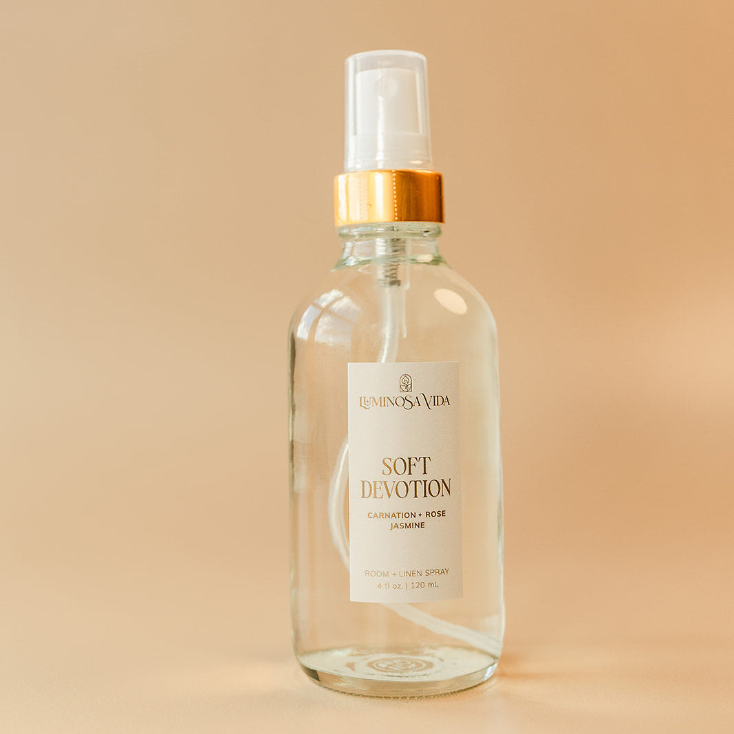 Luminosa Vida Soft Devotion room and linen spray with carnation, rose, and jasmine in a clear glass bottle with gold collar on a neutral background.