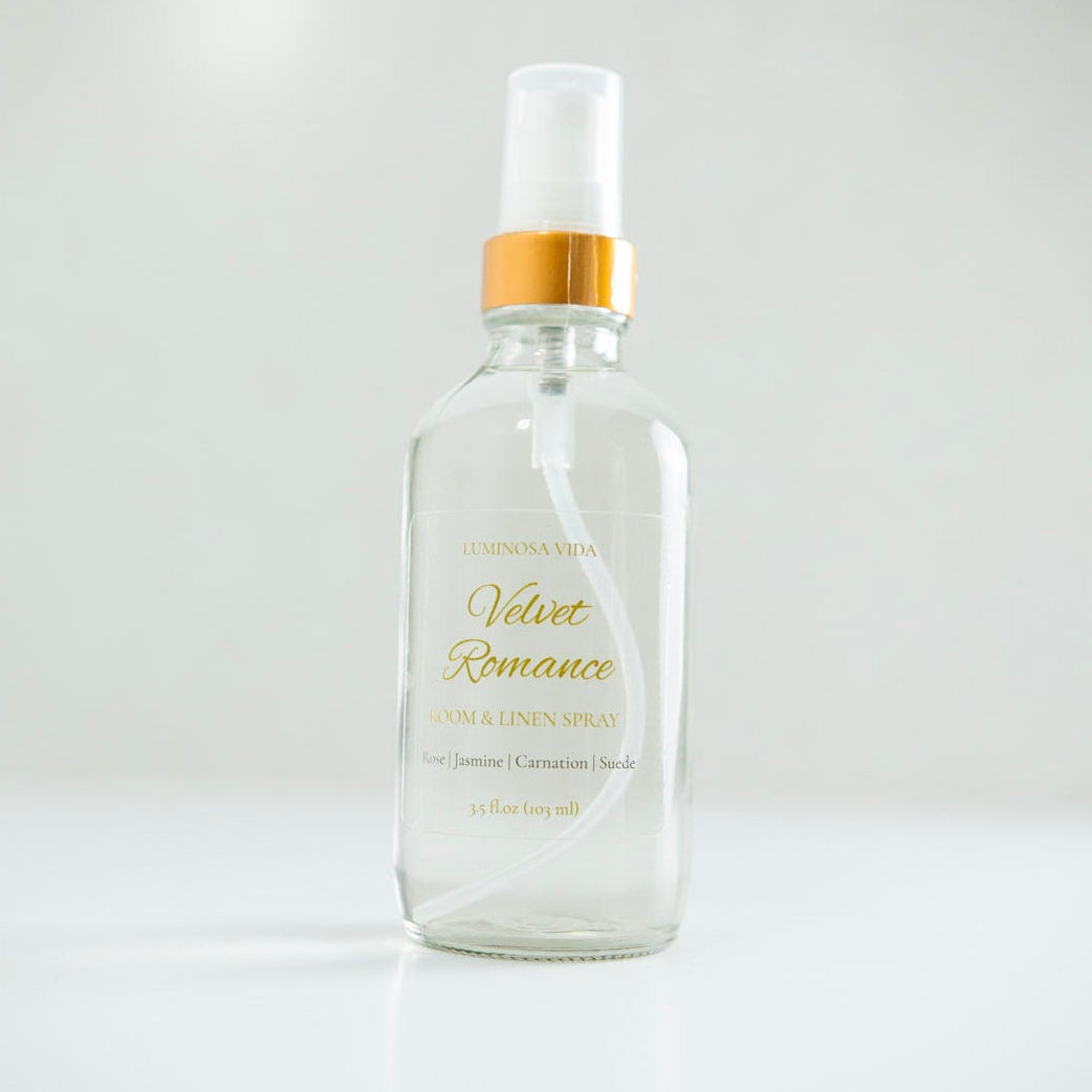“Velvet Romance Room + Linen Spray by Luminosa Vida displayed on a sleek white surface with a neutral background. A clear glass bottle with a gold cap and refined label featuring scent notes of Rose, Jasmine, Carnation, and Suede — a luxury, non-toxic home fragrance handcrafted in Austin, Texas.”
