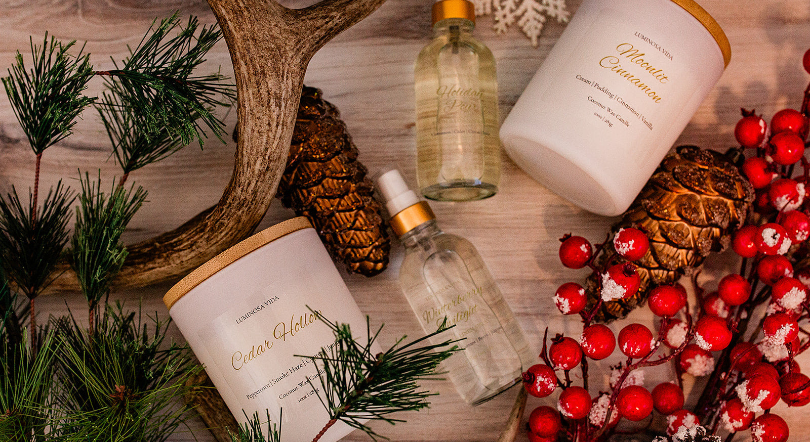 “Holiday flatlay featuring Luminosa Vida candles Cedar Hollow and Moonlit Cinnamon, accompanied by Winterberry Twilight and Velvet Amber room sprays. Styled with red berries, frosted pinecones, evergreen branches, and antlers on a rustic wooden background.”
