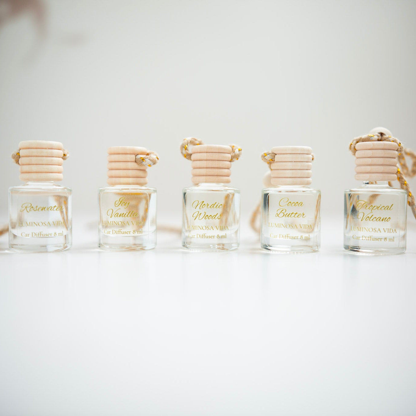 A row of seven elegant car diffusers from Luminosa Vida displayed on a clean white surface. Each small glass bottle features a light wooden cap with a coiled rope and gold text on the label. The scents shown are Dragon Fruit, Rosewater, Icy Vanilla, Nordic Woods, Cocoa Butter, Tropical Volcano, and Coconut Sandalwood.