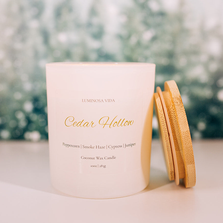 “Cedar Hollow candle by Luminosa Vida on a wooden surface with a winter forest background. White jar with a bamboo lid featuring notes of peppercorn, smoke haze, cypress, and juniper.”