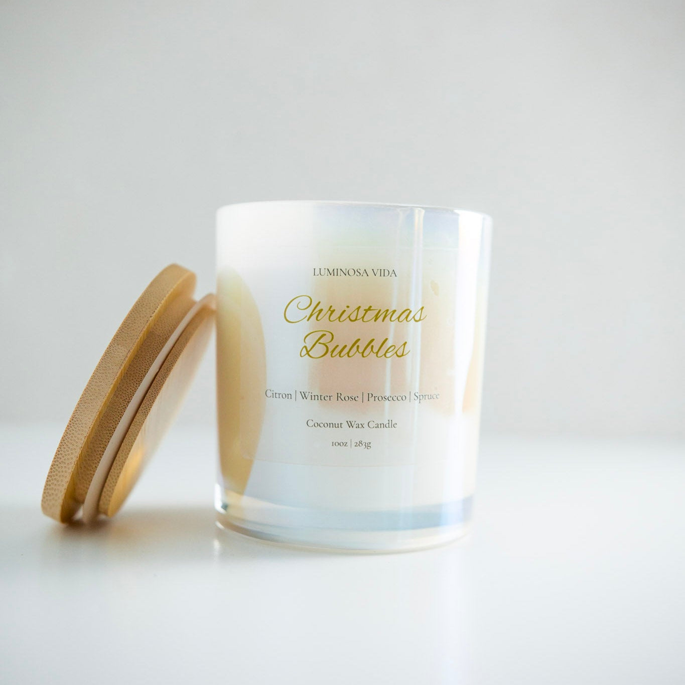 “Luminosa Vida Christmas Bubbles candle in an iridescent glass jar with a wood lid. Luxury coconut wax candle handcrafted in Austin, Texas with notes of citron, winter rose, prosecco, and spruce — a festive holiday scent.”