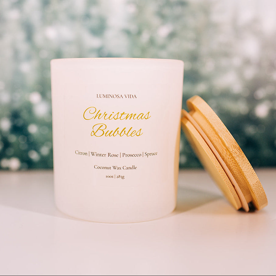 “Christmas Bubbles luxury holiday candle by Luminosa Vida on a wooden surface with a soft winter forest background. White jar with gold lid featuring notes of citron, winter rose, prosecco, and spruce.”