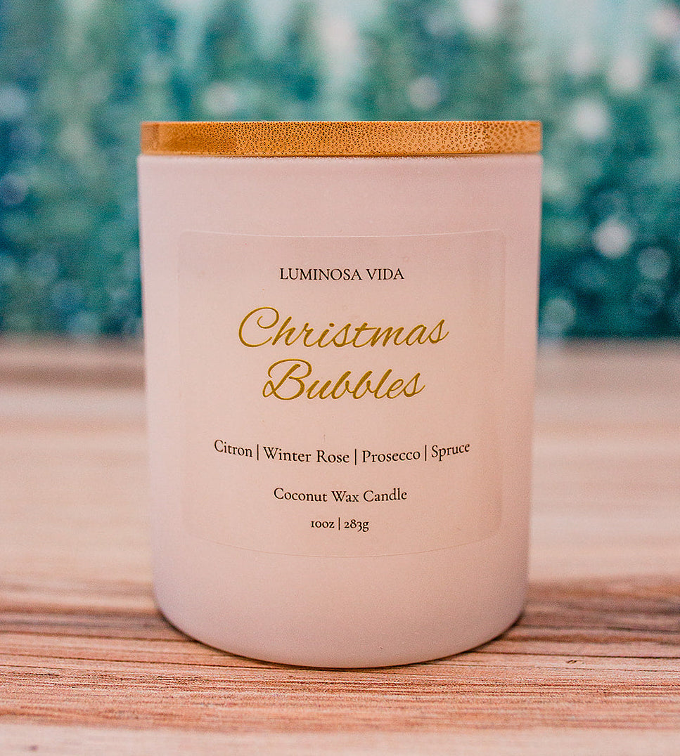 “Christmas Bubbles luxury holiday candle by Luminosa Vida on a wooden surface with a soft winter forest background. White jar with gold lid featuring notes of citron, winter rose, prosecco, and spruce.”