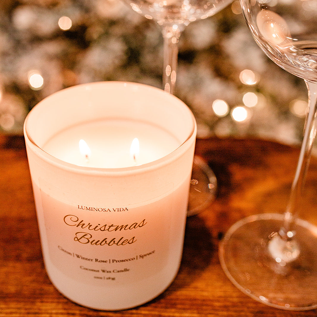 “Lit Christmas Bubbles candle by Luminosa Vida on a wooden surface with warm holiday bokeh lights and wine glasses in the background. White coconut wax candle featuring notes of citron, winter rose, prosecco, and spruce.”