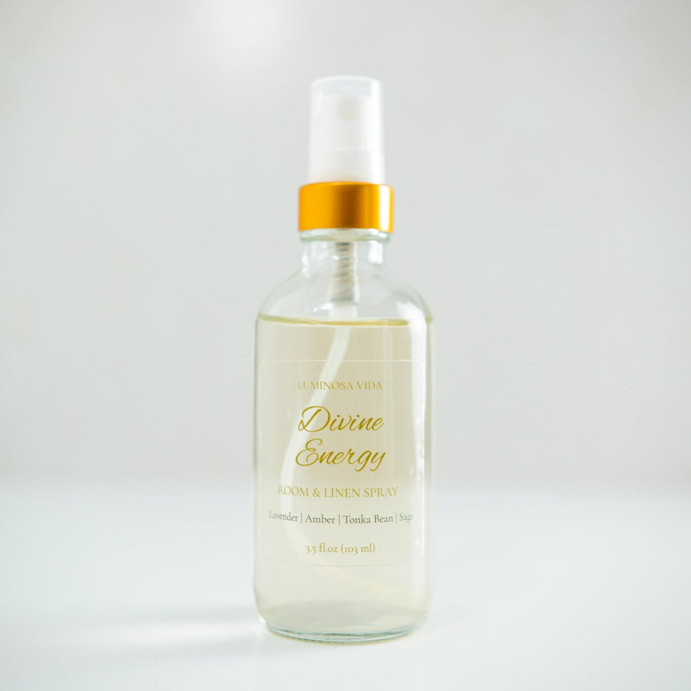 “Divine Energy Room + Linen Spray by Luminosa Vida displayed on a minimalist white surface with a soft neutral background. A clear glass bottle with a gold cap and elegant label featuring scent notes of Lavender, Amber, Tonka Bean, and Sage — a luxury, non-toxic home fragrance handcrafted in Austin, Texas.”