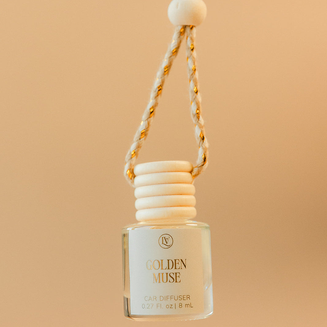 Luminosa Vida Golden Muse hanging car diffuser with braided cord in a warm neutral lifestyle setting.