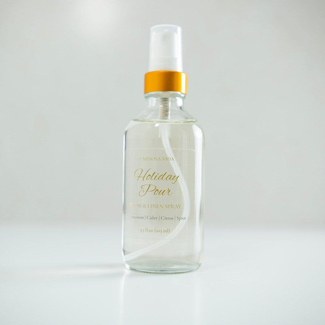 “Holiday Pour Room + Linen Spray by Luminosa Vida displayed on a minimalist white surface with a soft neutral background. A clear glass bottle with a gold cap and elegant label featuring scent notes of Cinnamon, Cider, Citrus, and Spice — a festive, non-toxic home fragrance handcrafted in Austin, Texas.”