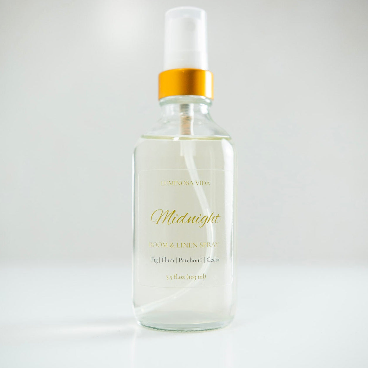 “Midnight Room + Linen Spray by Luminosa Vida displayed on a clean white surface with a soft gray background. A luxury clear glass bottle with a gold cap and elegant label featuring scent notes of Fig, Plum, Patchouli, and Cedar — a non-toxic home fragrance handcrafted in Austin, Texas.”