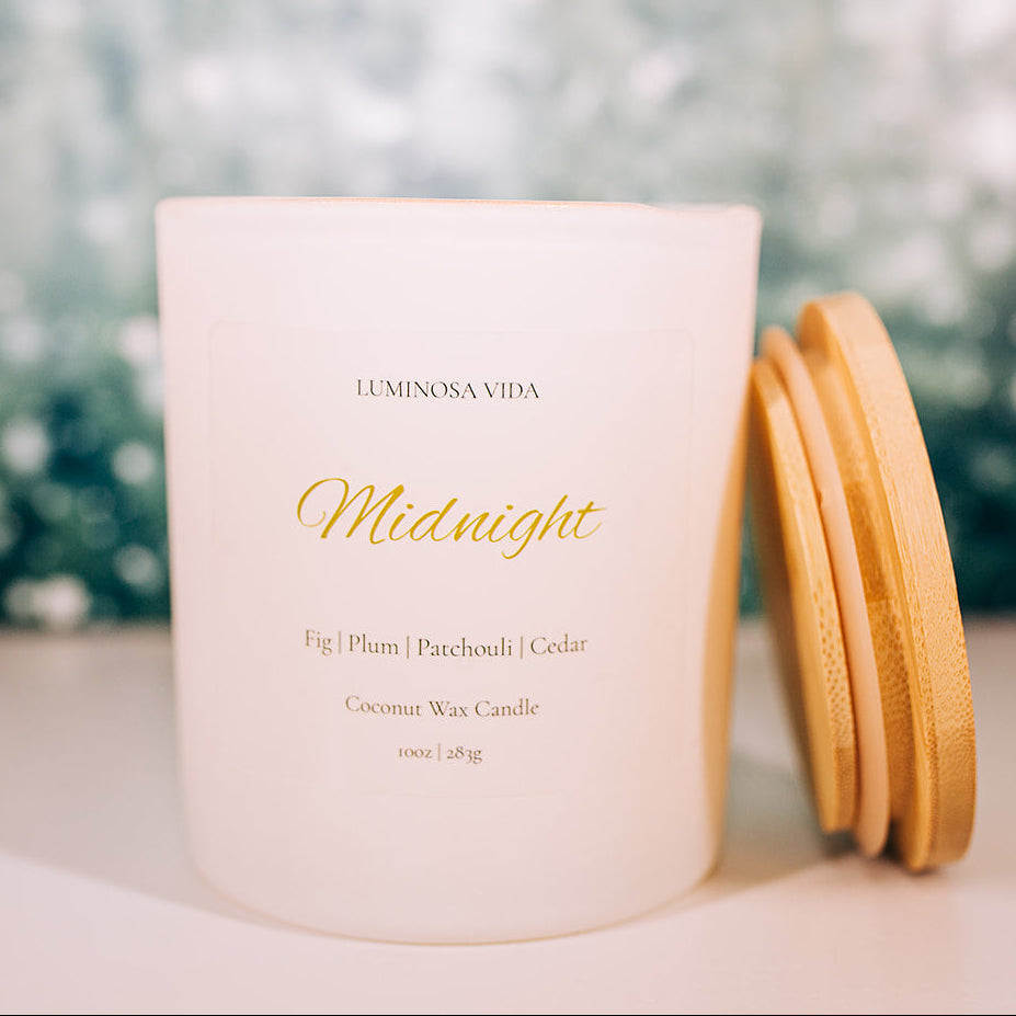 “Midnight candle by Luminosa Vida on a wooden surface with a soft winter forest background. White jar with bamboo lid featuring notes of fig, plum, patchouli, and cedar.”