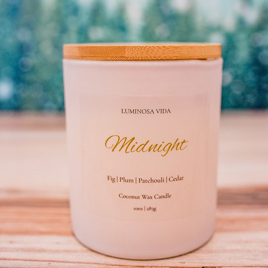 “Midnight candle by Luminosa Vida on a wooden surface with a soft winter forest background. White jar with bamboo lid featuring notes of fig, plum, patchouli, and cedar.”