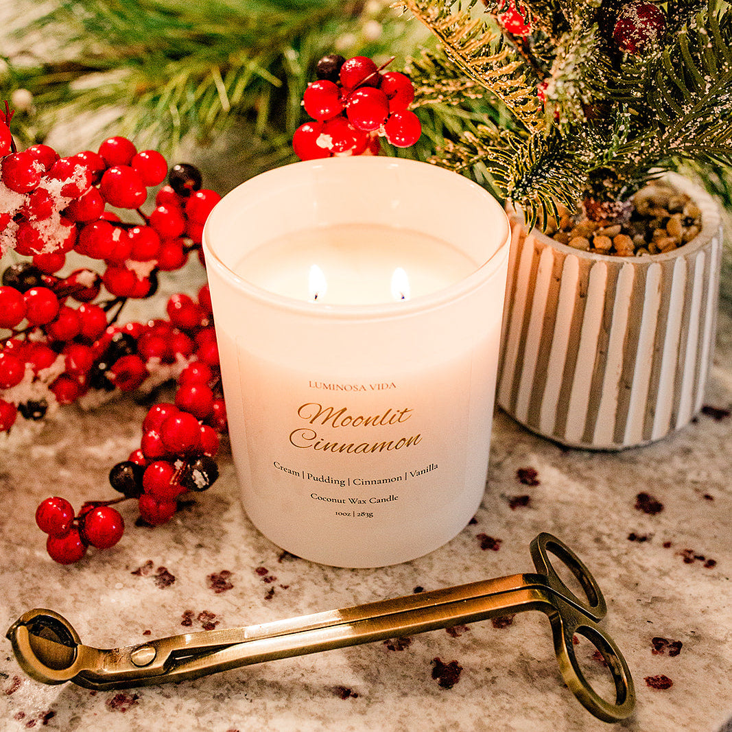 Lit Moonlit Cinnamon candle by Luminosa Vida displayed with frosted greenery and red berries, alongside a gold wick trimmer. White coconut wax candle featuring notes of cream, pudding, cinnamon, and vanilla