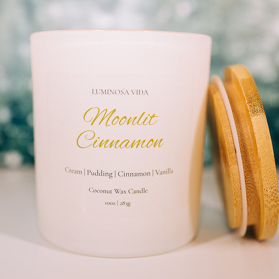 Moonlit Cinnamon candle by Luminosa Vida on a wooden surface with a snowy forest backdrop. White jar with bamboo lid featuring notes of cream, pudding, cinnamon, and vanilla.