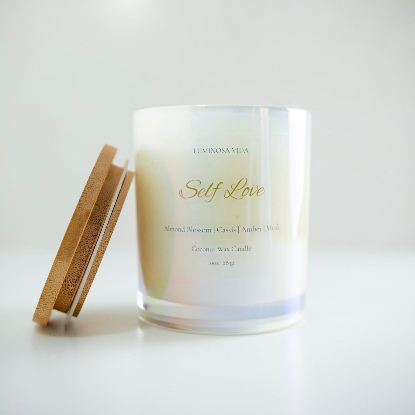 “Luminosa Vida Self Love candle in an iridescent glass jar with a wooden lid. Luxury coconut wax candle handcrafted in Austin, Texas with notes of almond blossom, cassis, amber, and musk — a warm, comforting scent that inspires self-care.”