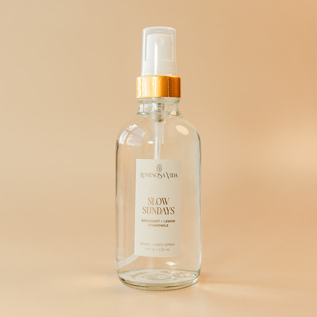 Luxury room and linen spray by Luminosa Vida in the Slow Sundays scent with bergamot, lemon, and chamomile in a clear glass bottle with gold collar on a soft neutral background.