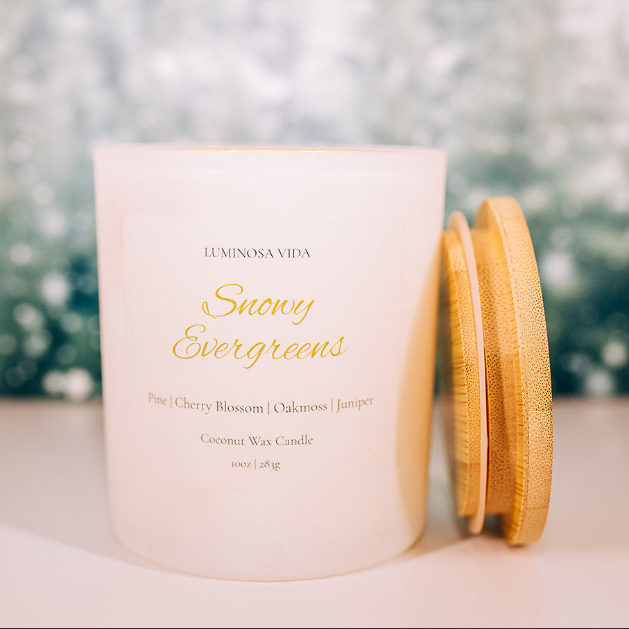 “Snowy Evergreens candle by Luminosa Vida on a wooden surface with a soft winter forest background. White jar with bamboo lid featuring notes of pine, cherry blossom, oakmoss, and juniper.”