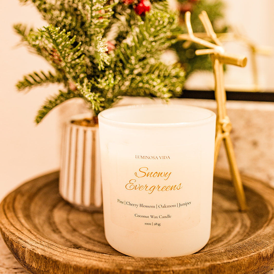 “Snowy Evergreens candle by Luminosa Vida displayed on a wooden tray with festive seasonal décor, including frosted greenery and a gold reindeer figurine. White coconut wax candle featuring notes of pine, cherry blossom, oakmoss, and juniper.”