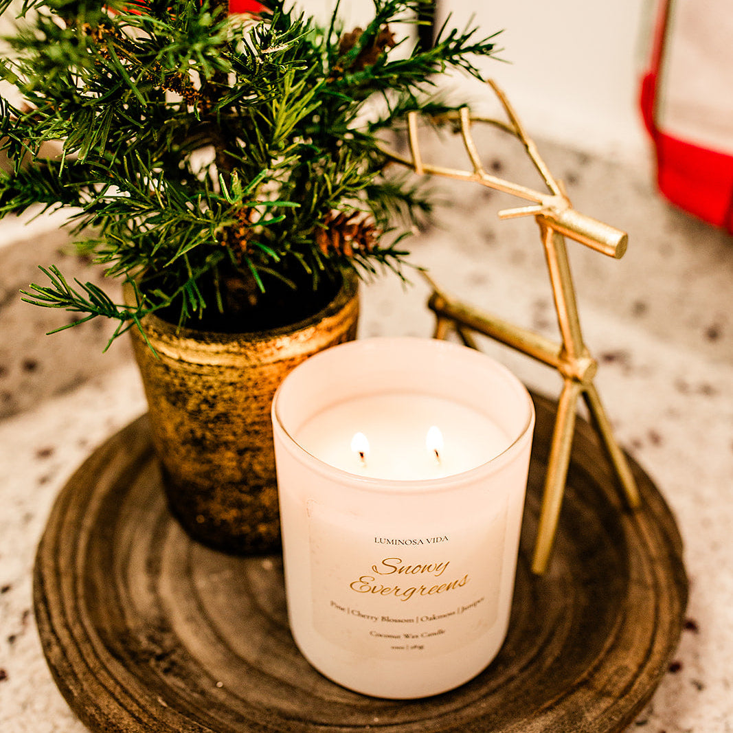 “Lit Snowy Evergreens candle by Luminosa Vida placed on a wooden tray with festive décor, including a small evergreen tree in a gold pot and a gold reindeer figurine. White coconut wax candle featuring notes of pine, cherry blossom, oakmoss, and juniper.”
