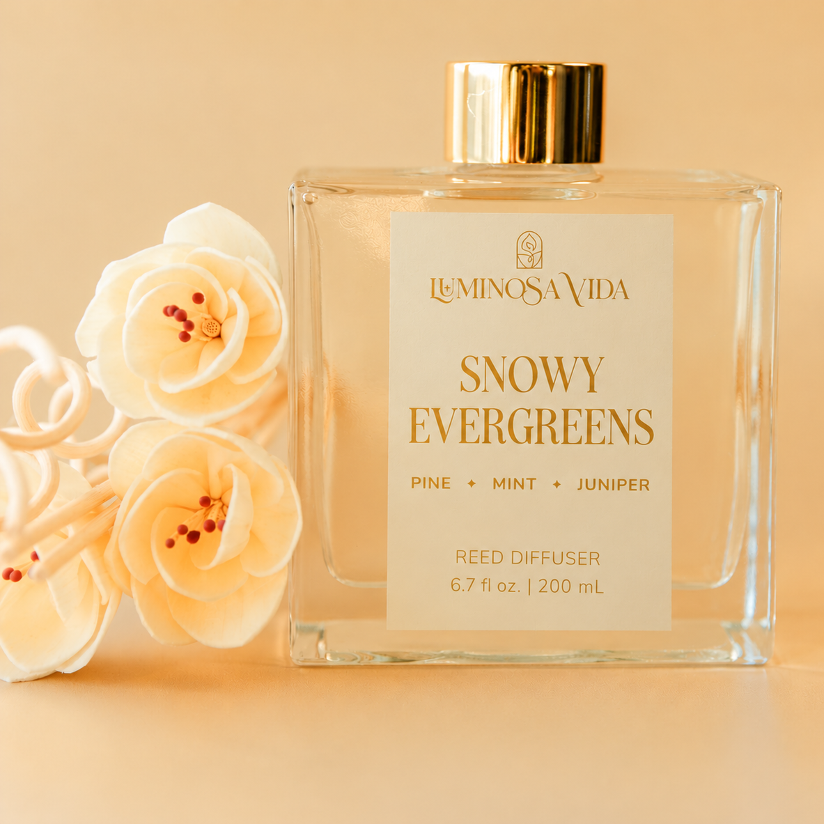 Luminosa Vida Snowy Evergreens Reed Diffuser in a clear glass bottle with gold cap, featuring pine, mint, and juniper scent notes against a warm beige background.